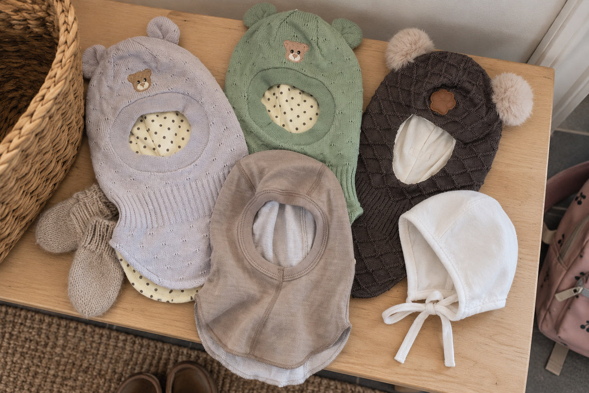 Balaclavaer til barn lagt ut på en benk: to Need it «Den Lune» i bomull med vindstopper, tynn ull/bambus-balaclava, tykk ullbalaclava med pote og en hvit babylue med knyting